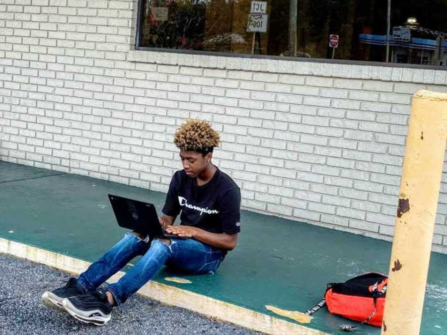 boy sits on curb outside of Subway with laptop completing schoolwork