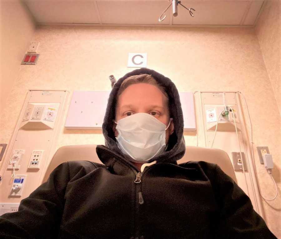 Blonde haired man wearing mask and black jacket in hospital room