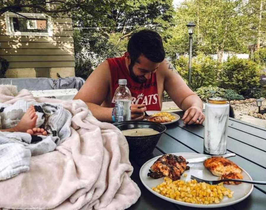 Foster dad eating chicken and corn next to newborn in carseat