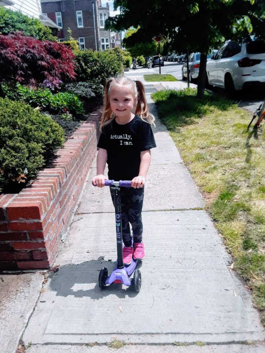 Little girl with Down syndrome riding scooter in "Actually, I can" shirt 