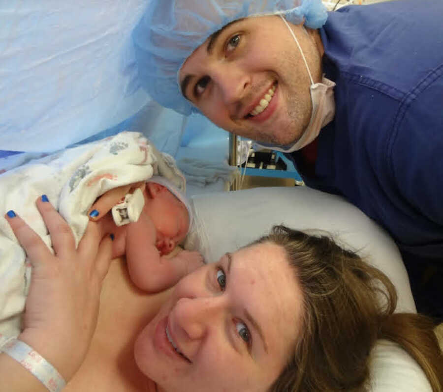 endometriosis warrior holds newborn baby on chest while husband stands behind her
