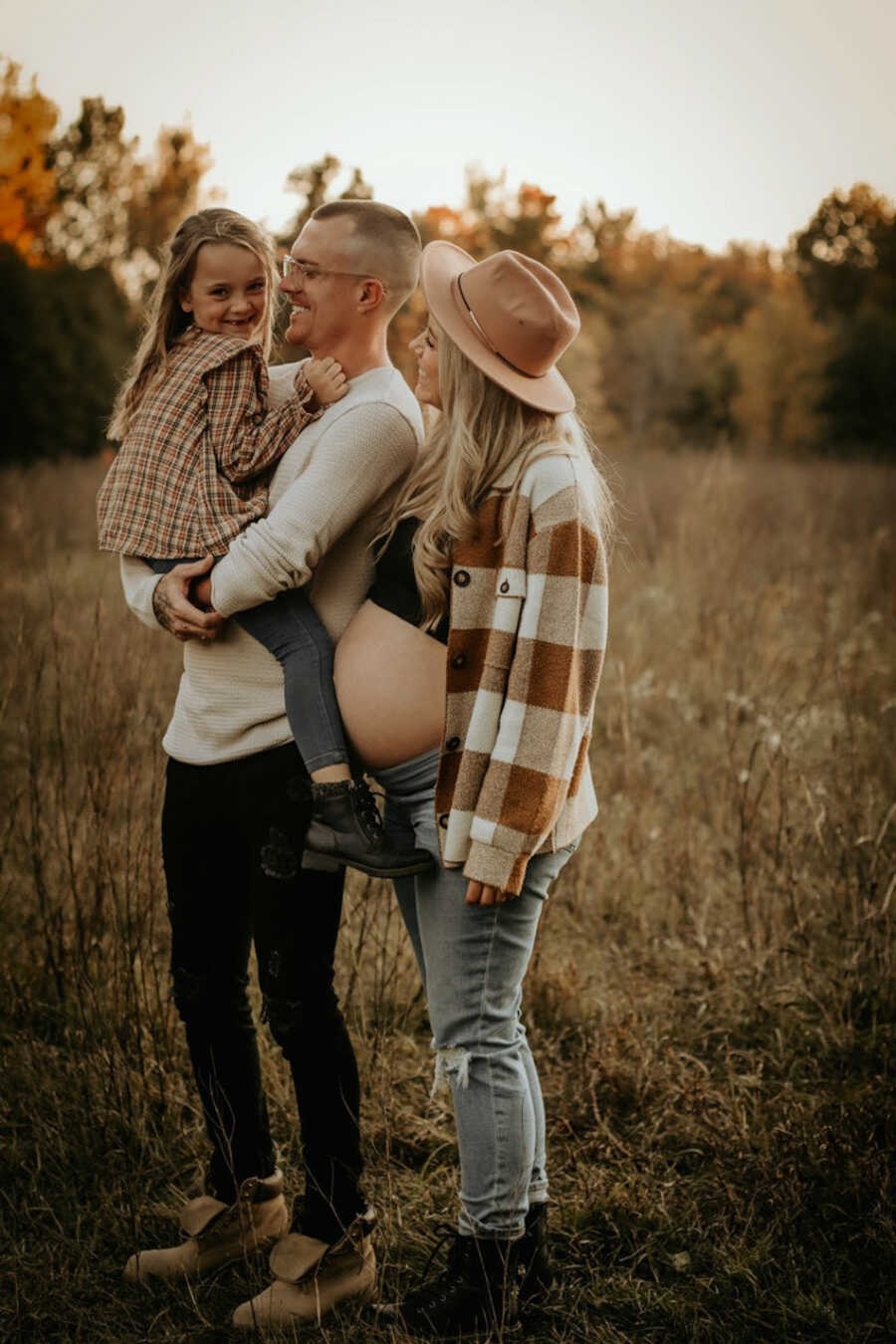 family photo of pregnant mom with husband holding daughter in a grass field