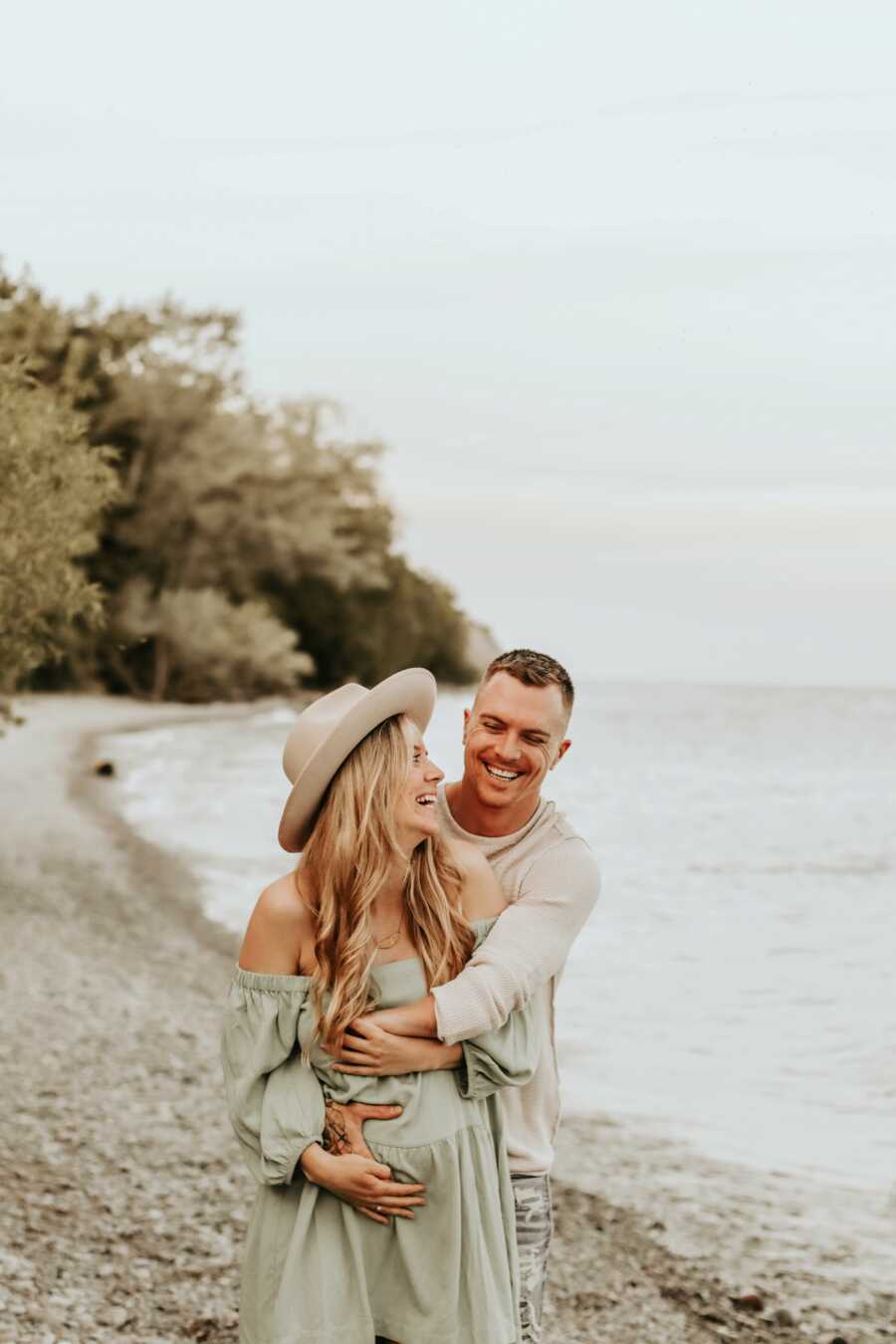 Pregnant mom wearing green dress and tan hat on beach with husband hugging her waist