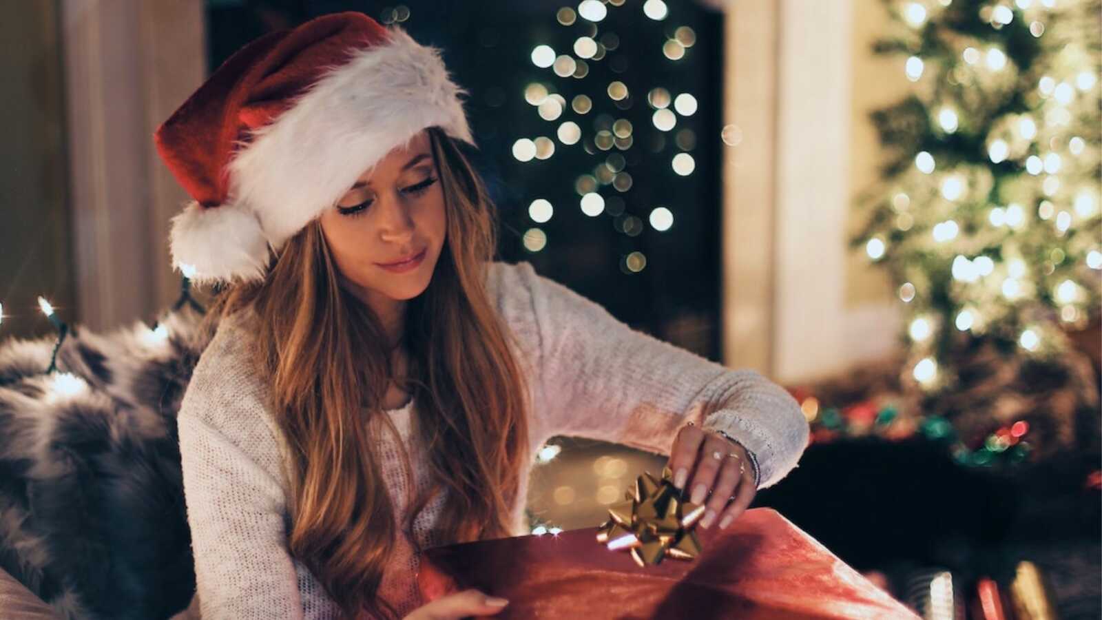 woman sits in front of Christmas tree in Santa hat, wrapping presents