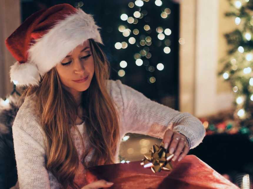 woman sits in front of Christmas tree in Santa hat, wrapping presents