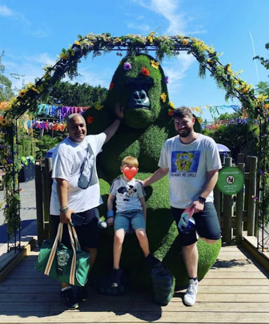gay couple stands with their adopted son in front of floral sculpture