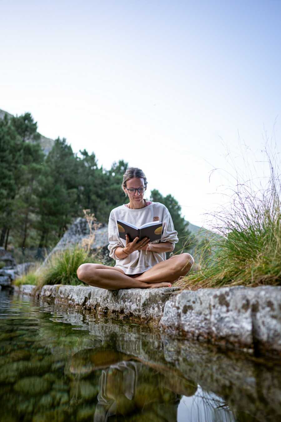 woman sits peacefully in nature by the water with a journal in hands