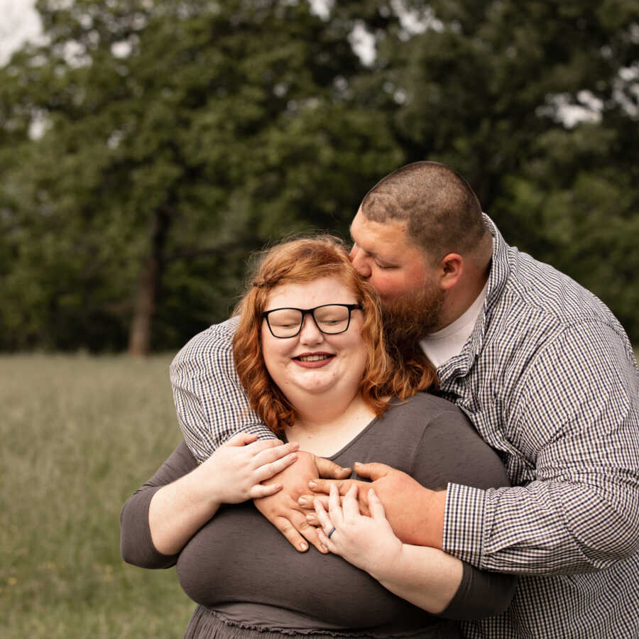 husband holds his wife from behind and gives her a kiss on the cheek
