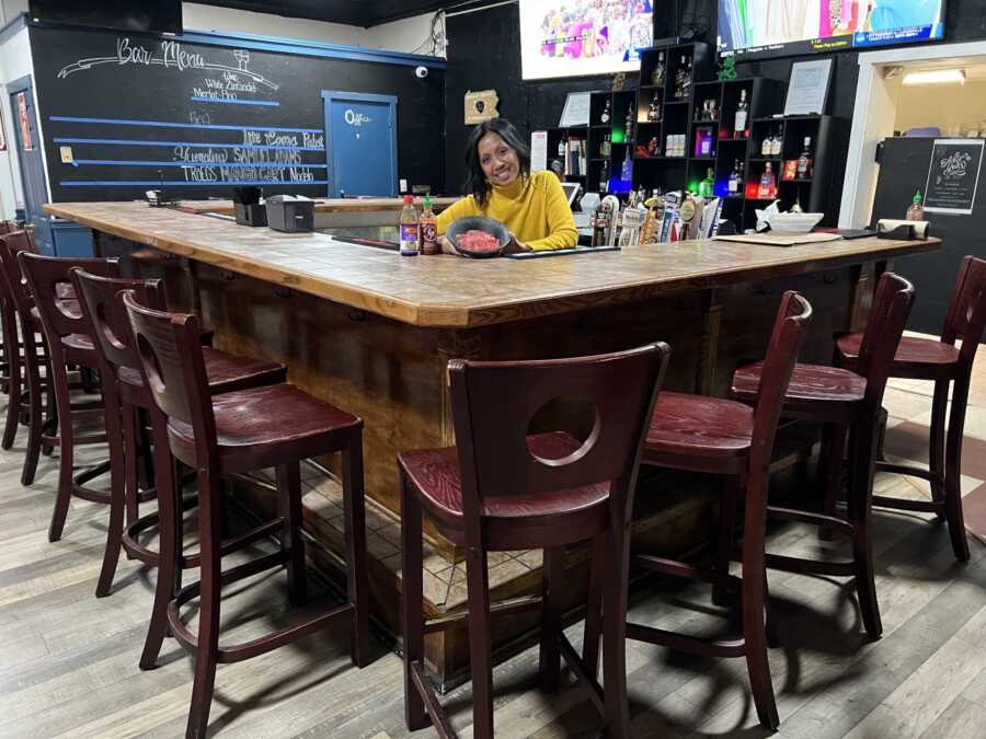 Small business owner of a bar standing behind the bar in a yellow shirt