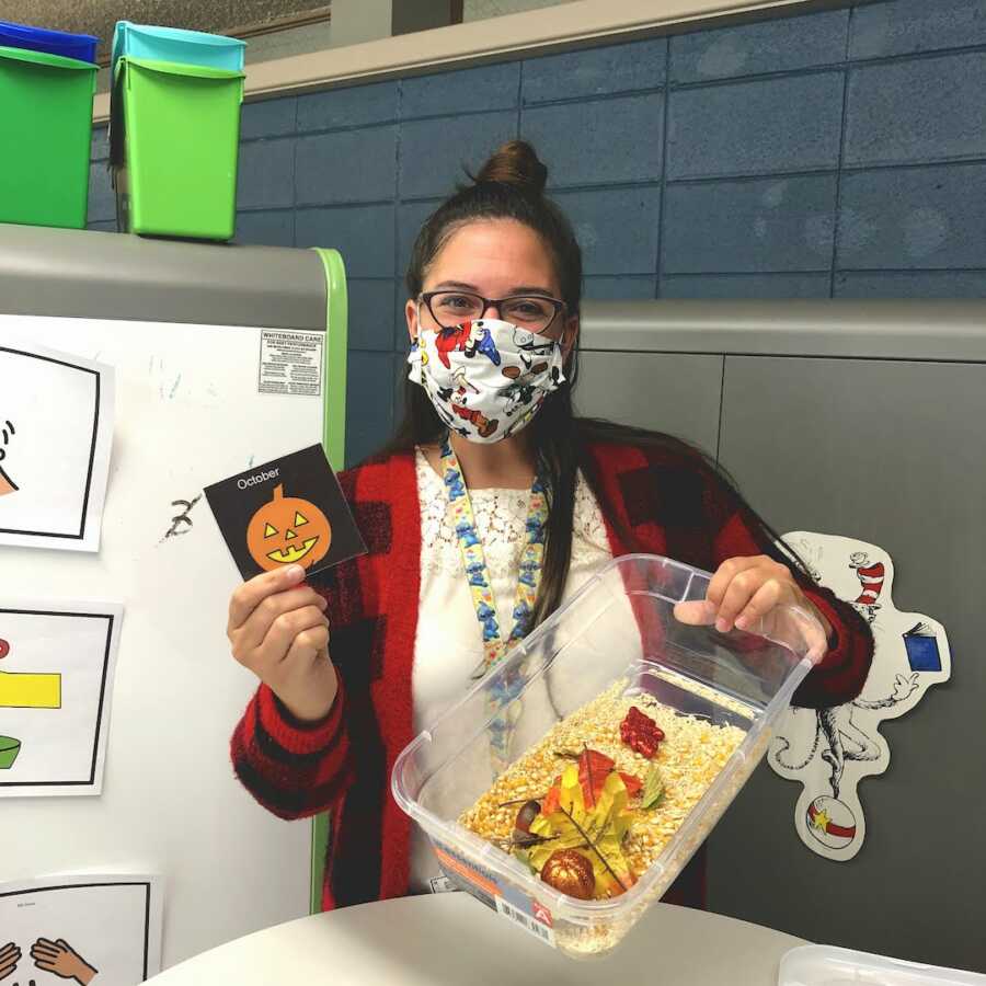 teacher holding up an "October" card and a bucket filled with sensory items