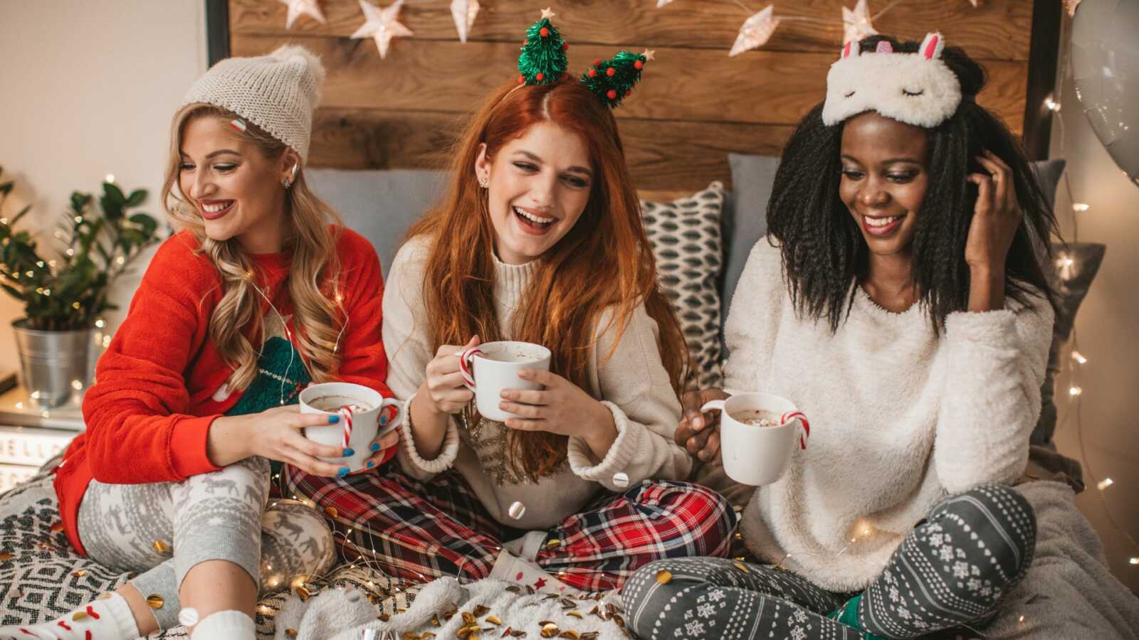three woman in festive outfits sit in bed drinking hot cocoa