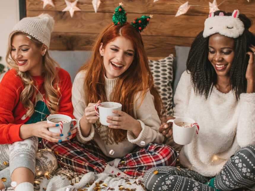 three woman in festive outfits sit in bed drinking hot cocoa