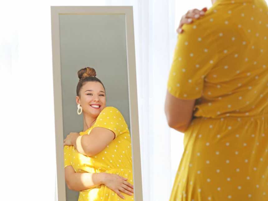 woman looks in mirror embracing herself and her body image