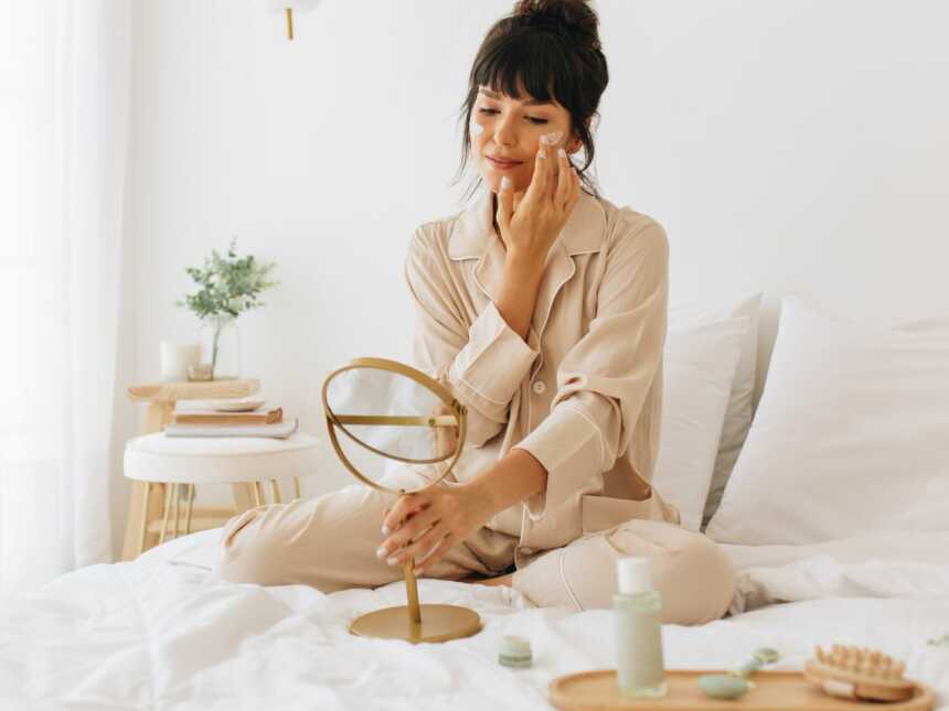 woman sits in bed applying skin care to her face as an act of self-care