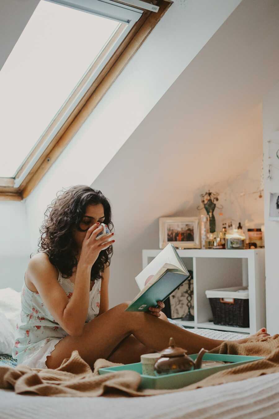 woman sits in bed sipping on tea and reading a book for self-care