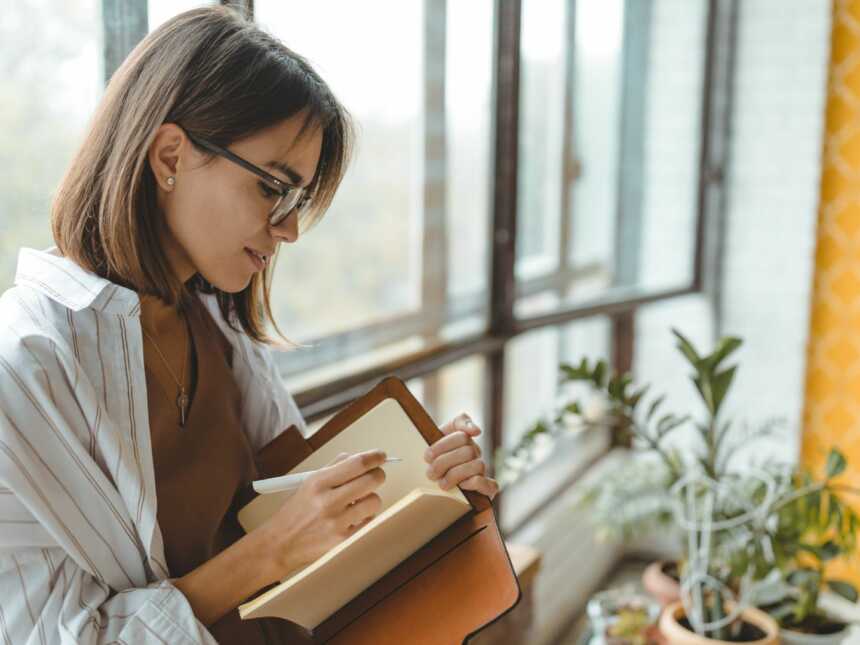woman sitting by window, wearing glasses, and journaling