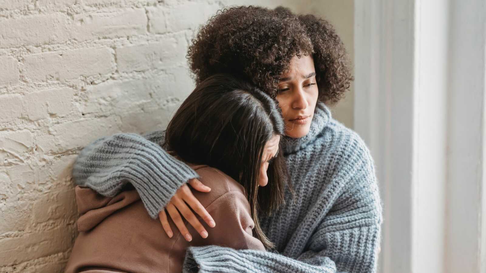 Woman hugs another woman against white brick wall