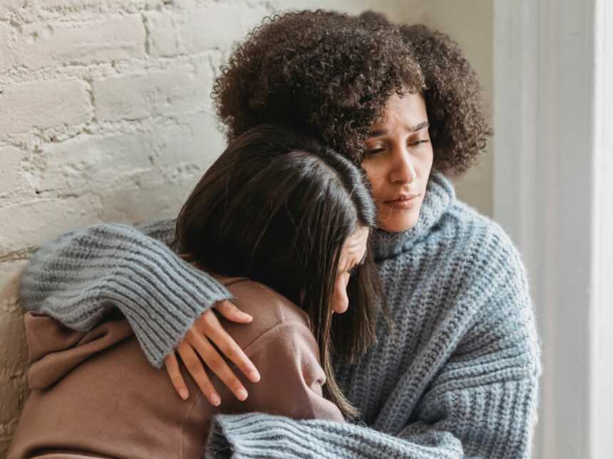 Woman hugs another woman against white brick wall