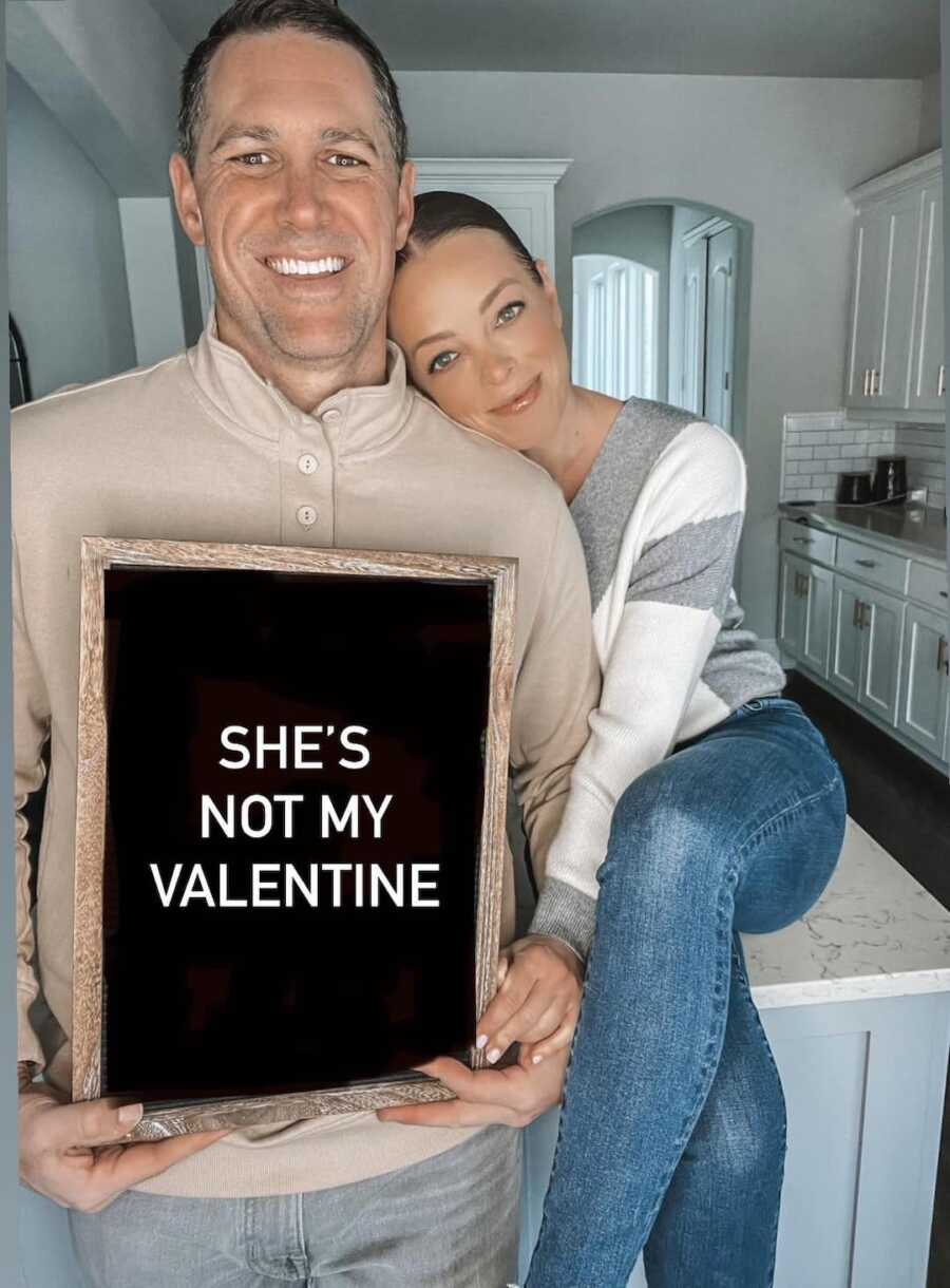 married couple standing in kitchen with husband holding sign reading "she's not my valentine"